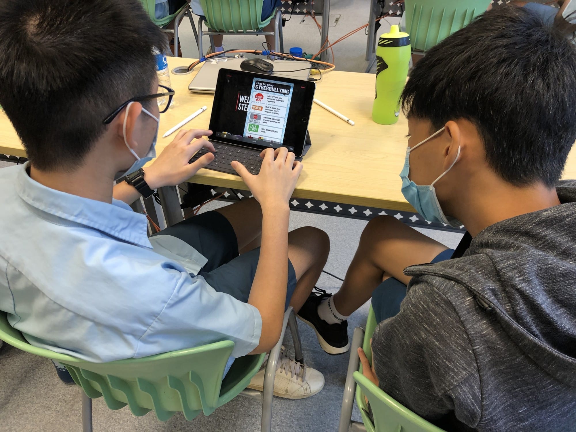 Two students sit side by side at a classroom table, using a tablet with a keyboard cover. Both wear face masks and look closely at the screen, which shows a poster titled “How to Stop Cyberbullying” with steps like stop, block, record, and tell someone. A stylus, a laptop, and a green water bottle rest on the desk, while light green chairs and cables are visible in the background.